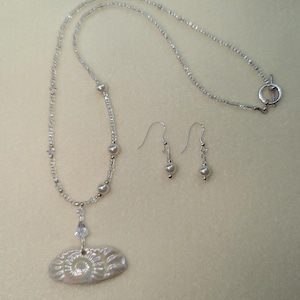 May include: A jewelry set consisting of a necklace and a pair of earrings. The necklace features a silver-toned chain with small clear beads, white pearls, and a central iridescent ammonite fossil pendant. The matching earrings have silver hooks.