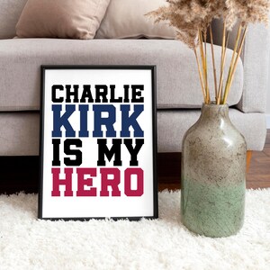 May include: Framed print with the text "CHARLIE KIRK IS MY HERO" in black, blue, black, and red lettering. The print is displayed on a white surface, with a vase of dried plants and a sofa in the background.