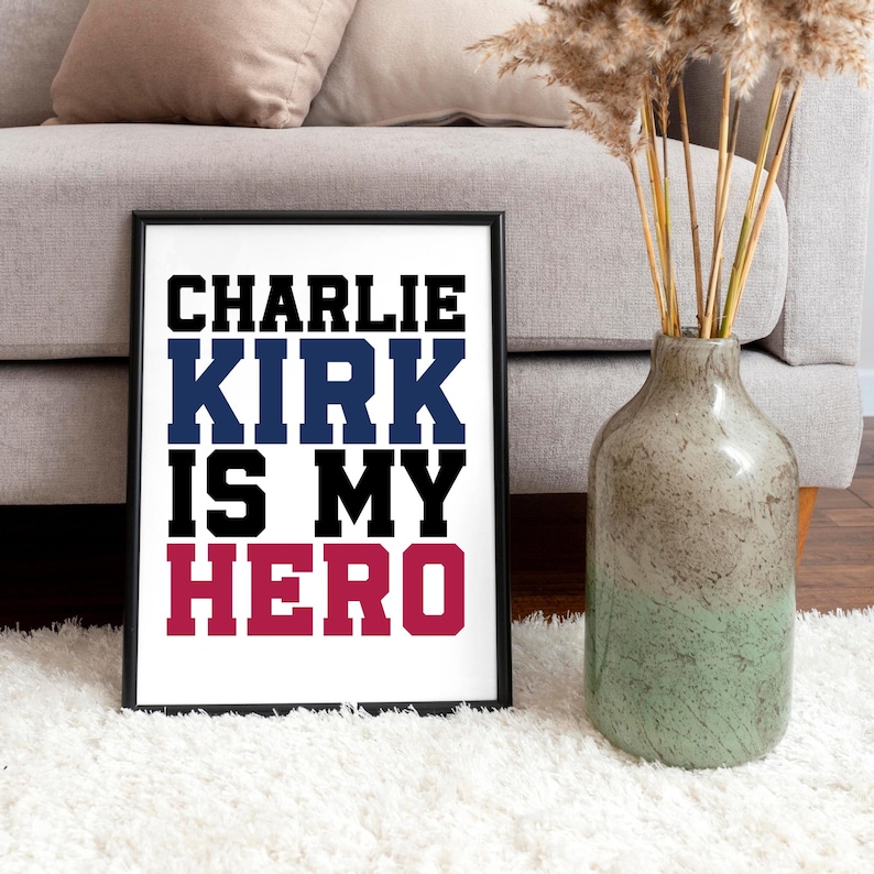 May include: Framed print with the text "CHARLIE KIRK IS MY HERO" in black, blue, black, and red lettering. The print is displayed on a white surface, with a vase of dried plants and a sofa in the background.