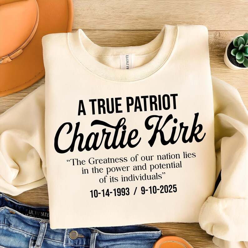 May include: Cream-colored sweatshirt with the text "A TRUE PATRIOT Charlie Kirk" in black. Below the name is a quote and dates. The sweatshirt is folded on top of blue jeans.