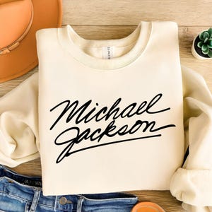 May include: Cream-colored sweatshirt with the name "Michael Jackson" in black cursive lettering. The sweatshirt is folded on a wooden surface, with a hat and jeans visible in the background. A small succulent plant is also present.