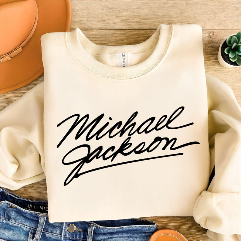 May include: Cream-colored sweatshirt with the name "Michael Jackson" in black cursive lettering. The sweatshirt is folded on a wooden surface, with a hat and jeans visible in the background. A small succulent plant is also present.