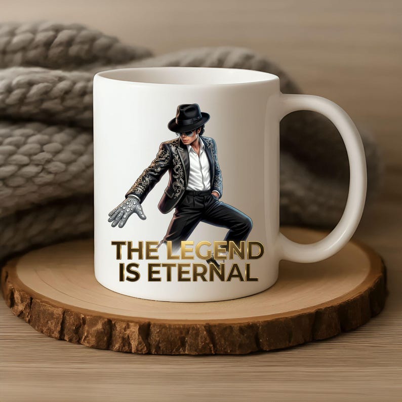 May include: White ceramic mug featuring an illustration of a man in a black suit and hat, with a silver glove. The text "THE LEGEND IS ETERNAL" is in gold. The mug is on a wooden coaster.