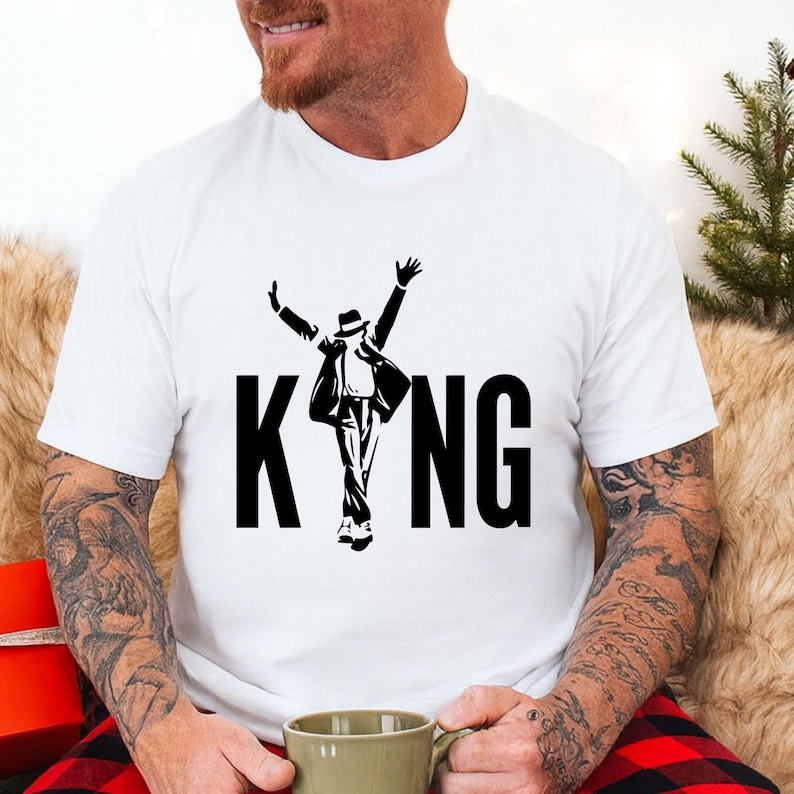 May include: White t-shirt featuring a black silhouette of a person in a hat and suit, with arms raised. The word "KING" is printed in bold, black capital letters below the figure. The shirt is made of a soft material.