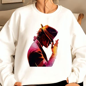 May include: White sweatshirt featuring a vibrant illustration of a person in a hat, with a purple jacket and a yellow glove. The artwork has a colorful, stylized design. The sweatshirt is a casual, comfortable garment.