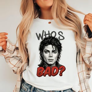May include: White t-shirt featuring a portrait of Michael Jackson with the text "WHO'S BAD?" in black and red. The graphic tee has a crew neck and short sleeves. The shirt is made of a soft, comfortable material.