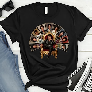 May include: Black t-shirt featuring a graphic of a man seated on a gold throne, surrounded by portraits. The central figure wears a black leather jacket and pants. The portraits are arranged in a semi-circle around the throne.