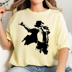 May include: A light yellow t-shirt featuring a black silhouette of a person in a hat, striking a pose. The graphic has a splattered paint effect. The shirt is made of a soft material.