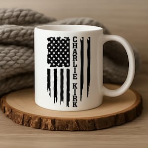 May include: White ceramic mug featuring a distressed American flag design with the text "CHARLIE KIRK" in black. The mug is set on a wooden coaster, with a soft, gray knitted blanket in the background.