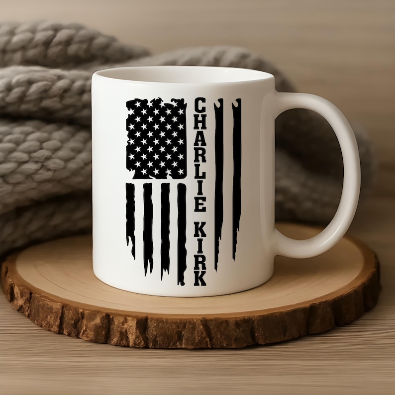 May include: White ceramic mug featuring a distressed American flag design with the text "CHARLIE KIRK" in black. The mug is set on a wooden coaster, with a soft, gray knitted blanket in the background.