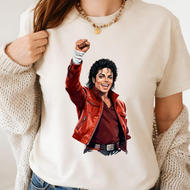 May include: Cream-colored t-shirt featuring a vibrant illustration of Michael Jackson in a red leather jacket, fist raised. The artwork captures his iconic look with a smiling expression. The shirt is a comfortable, casual wear.