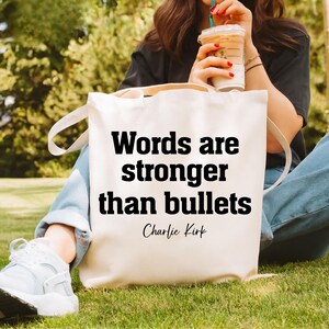 May include: Cream-colored tote bag with the quote "Words are stronger than bullets" in bold black letters. The quote is attributed to Charlie Kirk. The bag has white handles and is sitting on green grass.