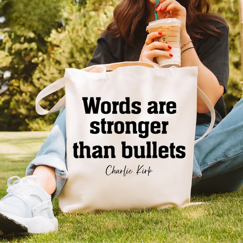 May include: Cream-colored tote bag with the quote "Words are stronger than bullets" in bold black letters. The quote is attributed to Charlie Kirk. The bag has white handles and is sitting on green grass.