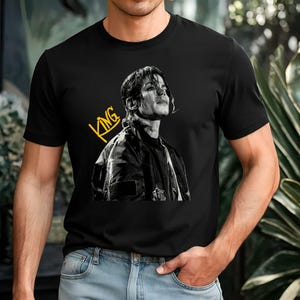 May include: Black t-shirt featuring a black and white portrait of a person with the word "KING" in yellow script. The person in the portrait is wearing a black jacket. The t-shirt is short-sleeved.