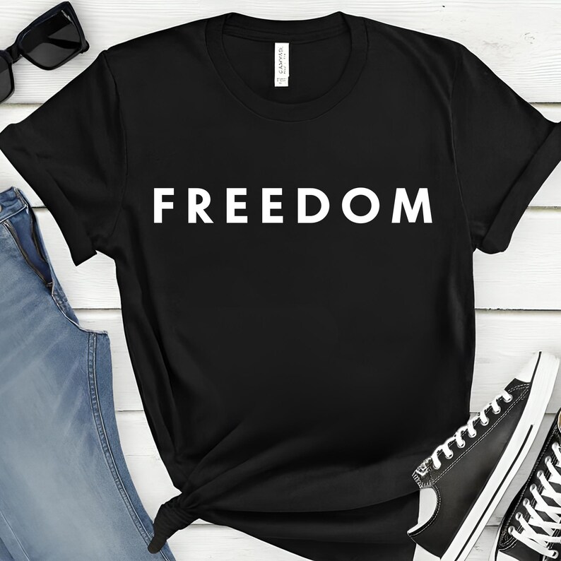 May include: Black t-shirt with the word "FREEDOM" printed in large, white capital letters. The shirt is paired with blue jeans and black sunglasses.
