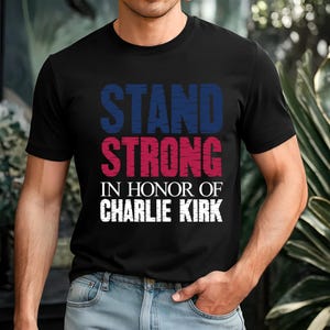 May include: Black t-shirt with the words "STAND STRONG" in blue and red, and "IN HONOR OF CHARLIE KIRK" in white. The shirt is a crew neck style.