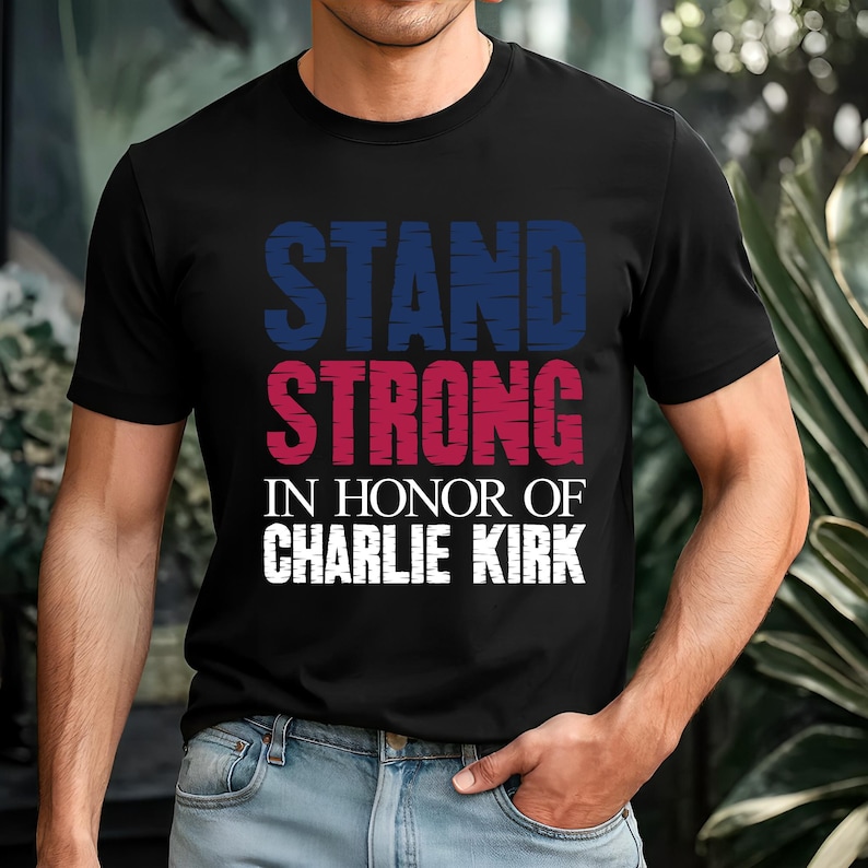 May include: Black t-shirt with the words "STAND STRONG" in blue and red, and "IN HONOR OF CHARLIE KIRK" in white. The shirt is a crew neck style.