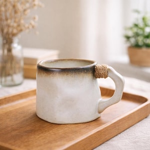 May include: A cream ceramic mug with a brown rim and a rope-wrapped handle. The mug is placed on a wooden tray. The mug has a simple, elegant design, suitable for tea or coffee. The mug's design suggests a rustic aesthetic.