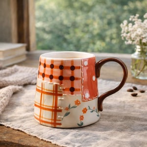 Pode incluir: Uma caneca de cerâmica com um design de patchwork. A caneca apresenta padrões de xadrez e bolinhas laranja, com uma alça marrom. Acentos florais são pintados na parte inferior da caneca. A caneca está cheia de um líquido escuro.