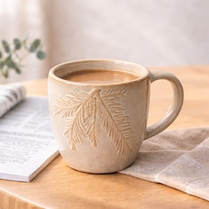 Puede incluir: Taza de cerámica beige llena de líquido marrón claro, posiblemente café o té. La taza presenta un diseño de hojas en relieve y un asa curvada. Se encuentra sobre una superficie de madera, junto a un paño doblado y un libro abierto.