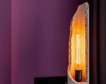 May include: A unique table lamp featuring a rough-edged, stone-like shade with a warm, glowing interior. The lamp has a long, exposed bulb and sits on a round, concrete base. The lamp is approximately 12 inches tall.