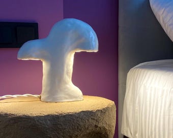 Puede incluir: Una lámpara blanca de cerámica con forma de hongo que emite un brillo suave, colocada sobre una mesa auxiliar texturizada de color marrón claro. El diseño de la lámpara es moderno y artístico, proyectando una luz cálida. El fondo presenta una pared púrpura y un vistazo de una cama.