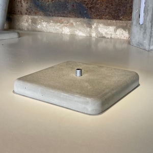 Può includere: Una base quadrata in cemento grigio chiaro con un inserto metallico filettato al centro. I bordi sono arrotondati. La base è probabilmente per una lampada o un altro oggetto decorativo.