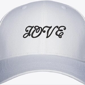 May include: A white baseball cap featuring the word LOVE embroidered in black thread across the front panel. The cap has a structured crown, a curved brim, and a button at the top centre. The stitching is clear and legible in a cursive font.