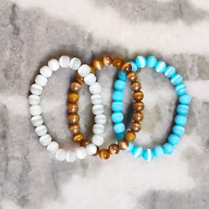 May include: Three beaded bracelets in white, brown, and blue. The bracelets are made of round beads and are arranged in a circular shape. The background is a light gray and white marble pattern.