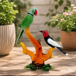 Puede incluir: Una escultura de cristal que muestra dos pájaros posados sobre una rama estilizada de color naranja y amarillo. Un pájaro es verde con el pico rojo, mientras que el otro es blanco y negro con la cabeza roja. La base es de cristal verde texturizado.