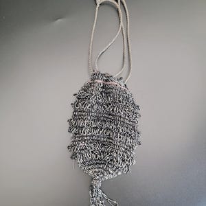 Può includere: Piccola borsa a coulisse di perline grigie e argentate. La borsa ha un corpo strutturato e frangiato e un nappa abbinata sul fondo. Un cordoncino argentato funge da coulisse e tracolla. Probabilmente vintage o fatto a mano.