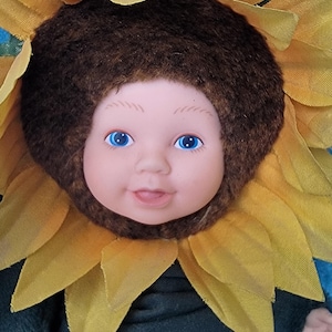 Puede incluir: Una muñeca bebé con un disfraz de girasol. La muñeca tiene cabello castaño, ojos azules y una tez rosada clara. El disfraz de girasol está hecho de tela amarilla con detalles en fieltro marrón.