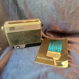 May include: A silver Kodak Instamatic M22 movie camera with a black instruction manual. The manual has the text "Kodak Instamatic M22 Movie Camera" printed on the front.