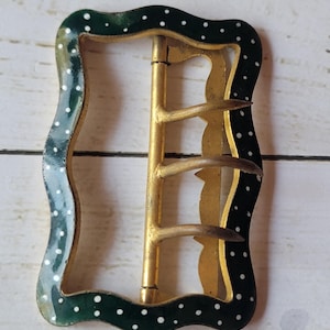 May include: An antique metal buckle with a gold-colored frame and a dark green, wavy border. The border is decorated with white polka dots. The buckle has three prongs and a pin.