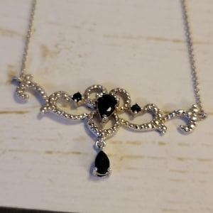 Op de afbeelding: Zilverkleurige ketting met een decoratief middelpunt met zwarte druppel- en ronde edelstenen. De ketting heeft een fijne ketting en is versierd met kleine, heldere kristallen.