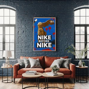 Puede incluir: Un póster enmarcado con el texto "NIKE BEFORE NIKE" en letras blancas sobre un fondo azul. La obra de arte presenta una estatua con alas y salpicaduras de pintura roja. El póster cuelga sobre un sofá color óxido.