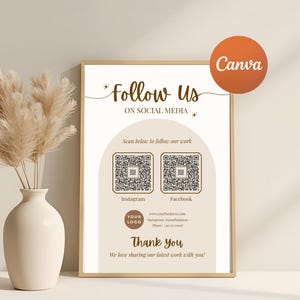 May include: A framed sign with the text "Follow Us on Social Media" and QR codes for Instagram and Facebook. The sign is in a beige color scheme with a gold frame. A vase with dried flowers is to the left.