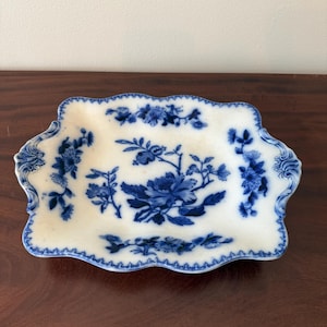 May include: A rectangular ceramic serving tray with scalloped edges and integrated handles, featuring a white base with a deep blue floral pattern. The design includes central peony-like blossoms and foliage, with decorative borders along the rim.