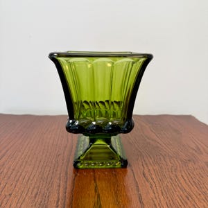 May include: A green glass vase with a square base and a flared, fluted top. The vase has a dark green rim and base, and the glass has a ribbed design. The vase is sitting on a wooden surface.