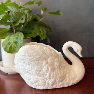May include: A white ceramic swan-shaped planter featuring detailed molded feather textures across its body, a curved neck, and an open top cavity for holding items. The swan is finished in a matte white glaze with a smooth, clean surface appearance.
