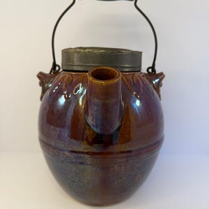 May include: A vintage brown ceramic teapot with a metal lid and handle. The teapot has a rounded body, a spout, and two small decorative handles. The handle is black and the lid is a silver color.