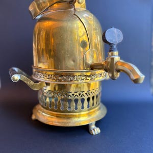 May include: A vintage brass tea or coffee pot with a spigot and a handle. The pot has a decorative base with a patterned design and small feet. The top has a hinged lid and a handle. The pot is made of brass and has a shiny finish.