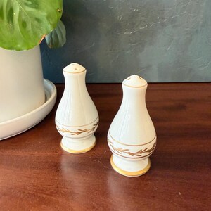 May include: A pair of white ceramic salt and pepper shakers with a tapered, bulbous shape. Each shaker features a delicate gold-colored leaf pattern encircling the lower body, with gold trim along the base and the top rim of the shaker lid.
