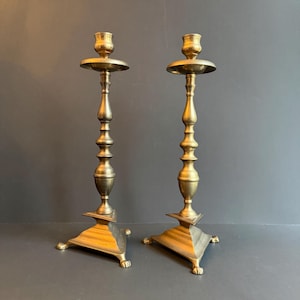Puede incluir: Dos candelabros antiguos de latón. Cada candelabro tiene una base cuadrada con patas de garra, un tallo detallado y una parte superior circular para una vela. Los candelabros son de color dorado cálido.