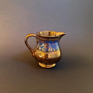 May include: A small, decorative creamer with a gold-colored exterior and a blue interior. The creamer has a handle and a spout for pouring. The design includes gold accents and a detailed pattern around the rim. The creamer is approximately 4 inches tall.