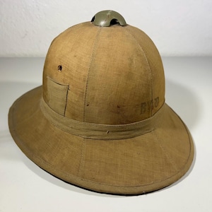 Può includere: Un elmetto coloniale beige con tesa larga e corona arrotondata. Il cappello presenta un piccolo sfiato verde oliva sulla parte superiore e una fascia attorno alla base della corona. Una piccola toppa quadrata è sul lato. Il cappello ha le lettere "BVJ" sul lato.