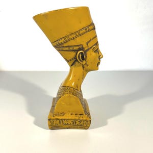 Könnte beinhalten: Eine senfgelbe Büste der Nofretete, der Großen Königlichen Gemahlin des ägyptischen Pharaos Echnaton. Die Büste zeigt eine hohe, flachköpfige Krone, einen schlanken Hals und detaillierte Gesichtszüge. Der Sockel hat hieroglyphische Schnitzereien.