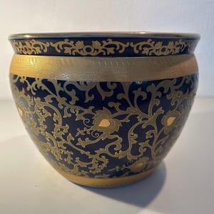 Könnte beinhalten: Ein tief dunkelblauer Keramikübertopf mit einem komplizierten goldenen Filigranmuster aus geschwungenen Ranken und Blättern. Das schalenförmige Gefäß hat ein breites, glattes Goldband am oberen Rand und einen massiven goldenen Boden.
