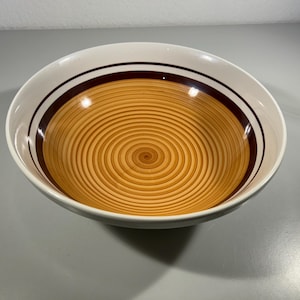 Könnte beinhalten: Eine weiße Keramikschale mit einem braunen und beigen Spiraldesign. Die Schale hat einen breiten Rand mit einem dunkelbraunen Band und einem helleren braunen Band. Das Innere der Schale weist ein Spiralmuster in Beige- und Brauntönen auf.
