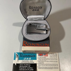 May include: A silver-toned Ronson gas lighter rests inside a dark grey, oval-shaped presentation case with a white satin lining. The lighter sits atop its original cardboard box, accompanied by a printed instruction manual featuring Ronson branding.